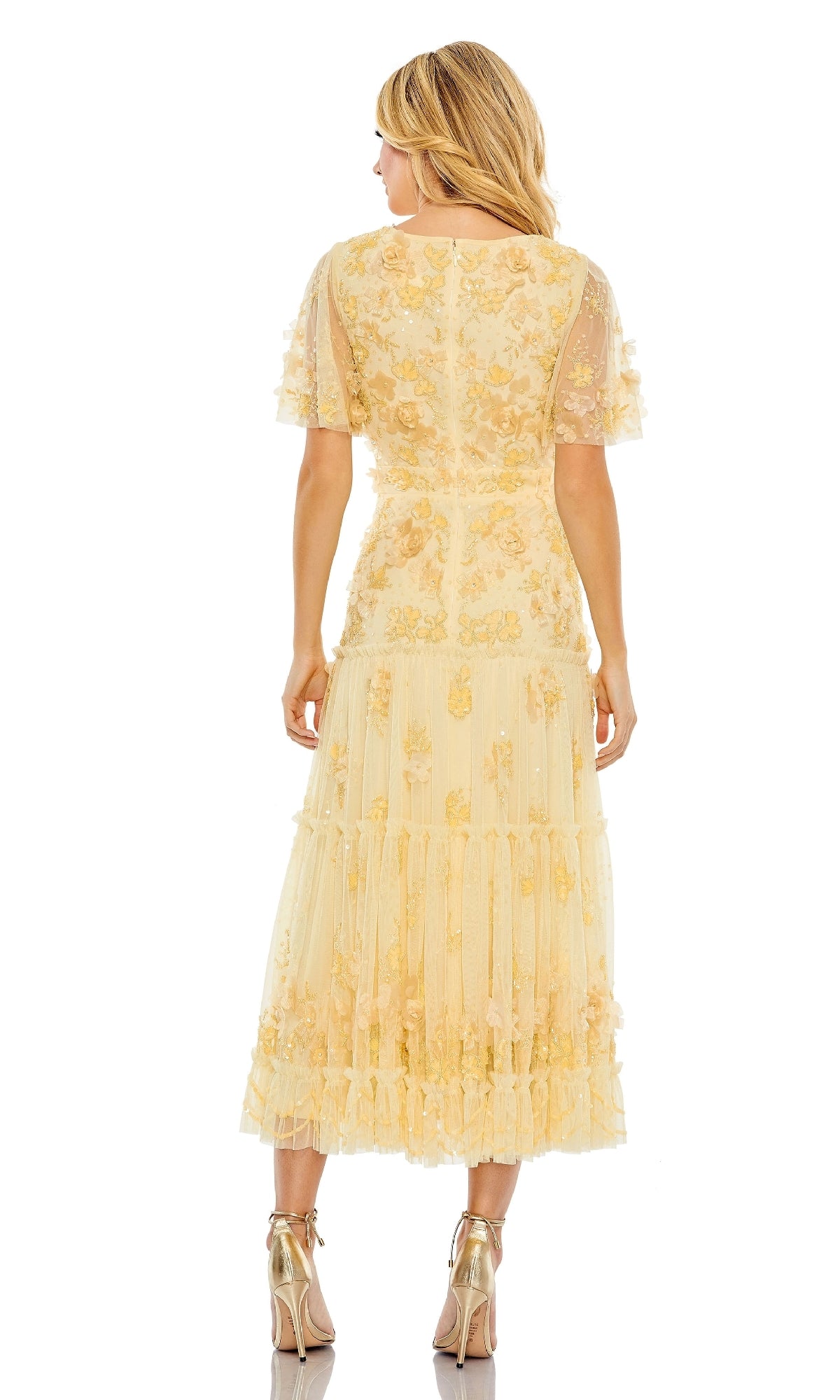 A woman faces away, wearing the Mac Duggal 9207 A-Line Wedding Guest Dress—light yellow, floral-embellished, with short sleeves and a tiered skirt—paired with metallic high-heeled sandals against a white background.